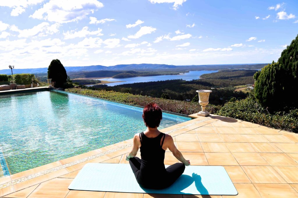 Understand what seasonal affective disorder is and how to manage symptoms. A brown-haired woman is seated in a yoga position facing very strong sunshine amidst mountains and valleys.