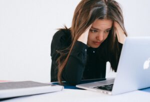 A young woman stares stressfully at a computer screen with her hands holding her head.