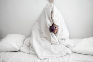 Several contributing factors may lead to seasonal affective disorder. A person hiding under a large doona holding a hot cup of tea trying to escape the cold.